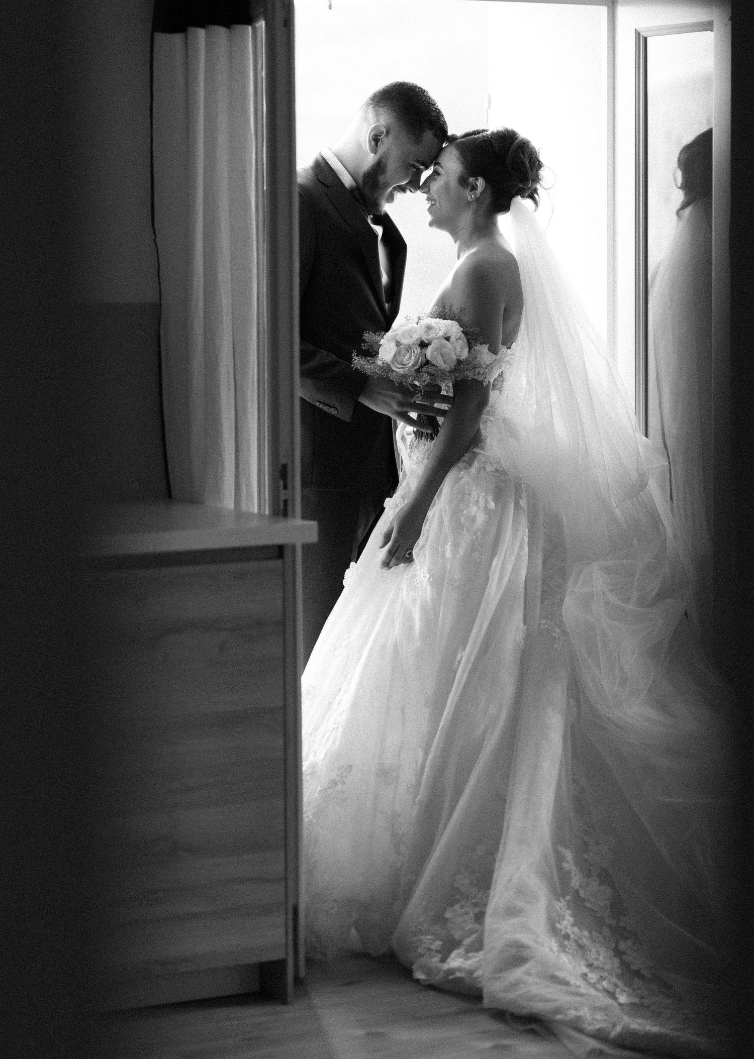 Black and white silhouette of bride and groom sharing intimate moment in doorway with veil flowing.