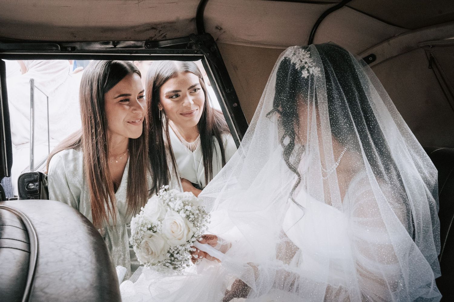 Bride in white gown and veil sits in vintage car with bridesmaids holding white bouquet.