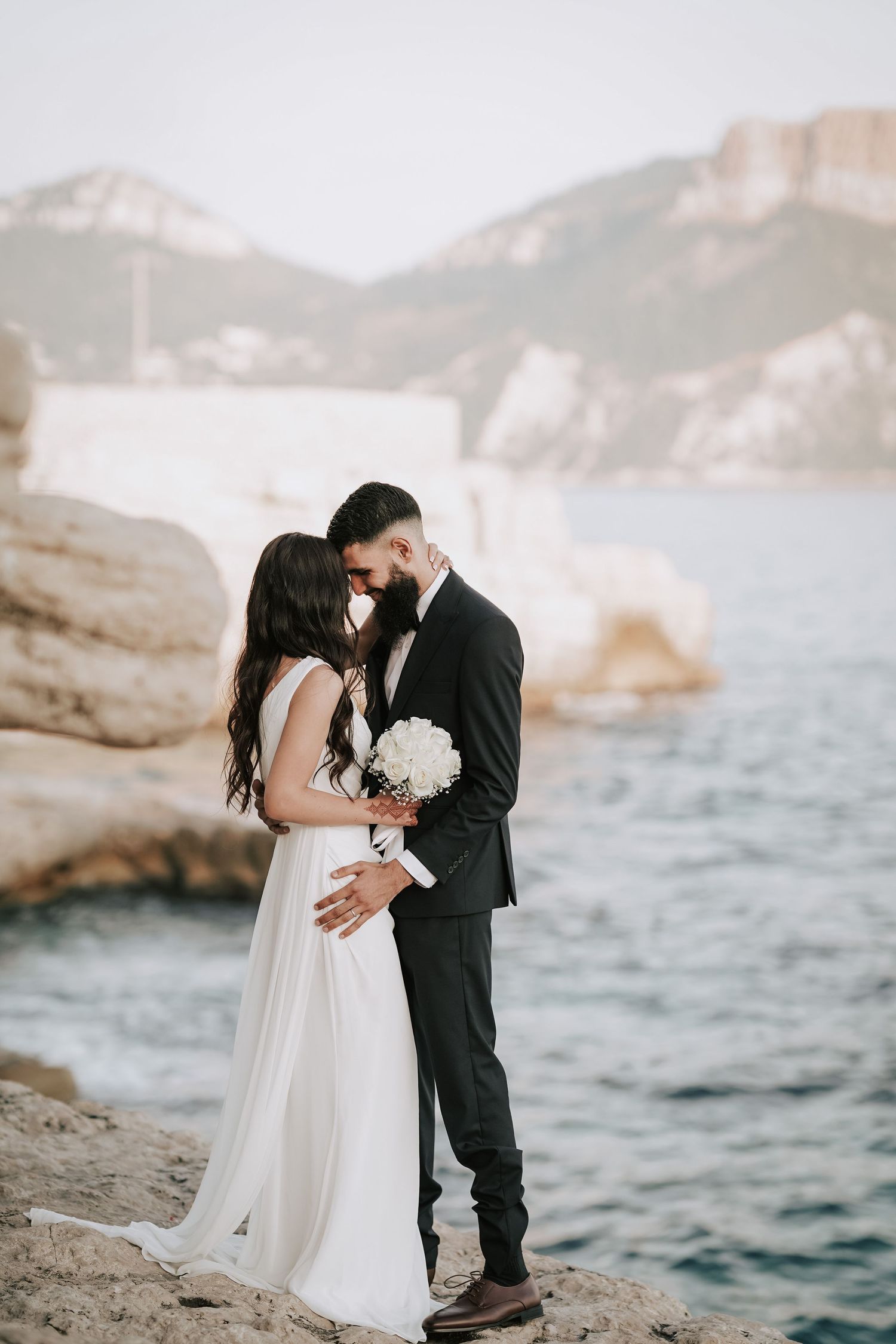 Bride and groom share a tender kiss on coastal rocks with mountains and sea in the background.