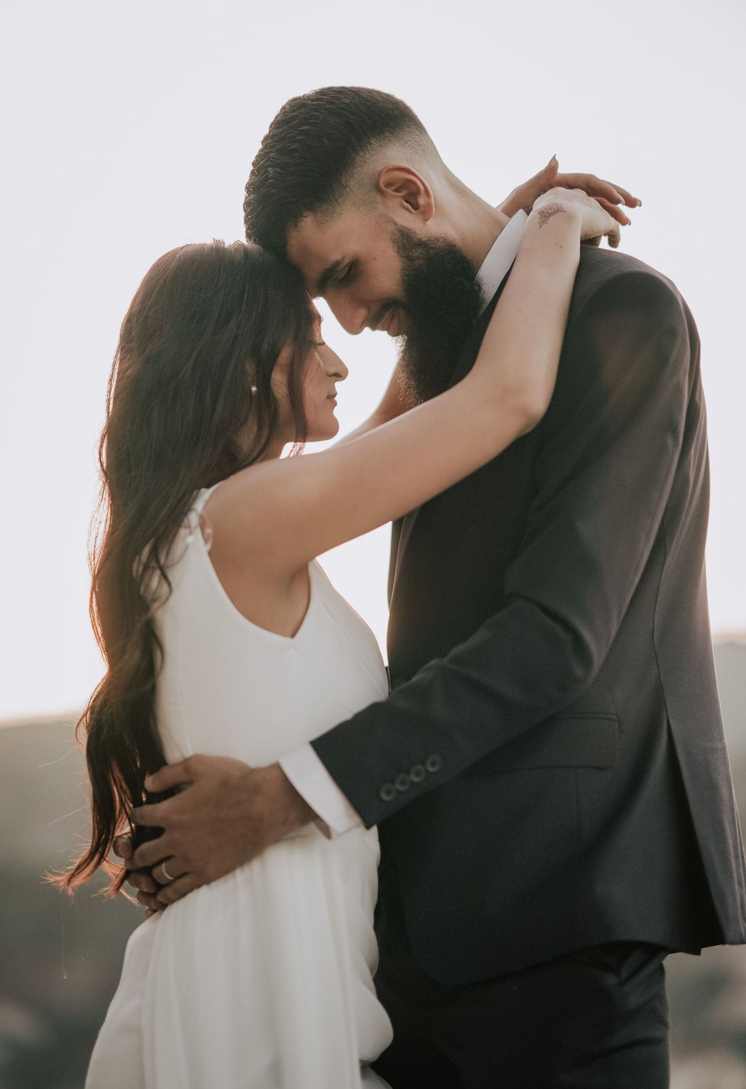 Bride and bearded groom share an intimate embrace, foreheads touching, in a romantic outdoor setting.