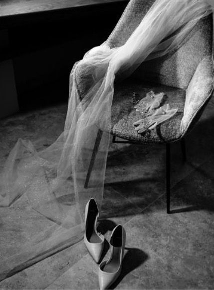 Black and white photo of a bridal veil draped over a chair beside elegant pointed-toe heels on a concrete floor.