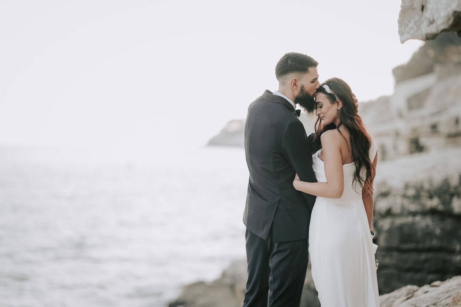 Bride and groom stand face-to-face on rocky cliffs with the ocean glowing softly behind them.