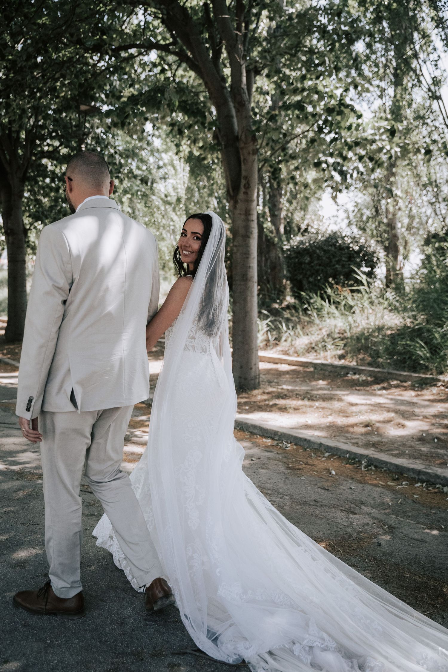 Bride in a flowing white gown and veil walks with groom through a tree-lined path.