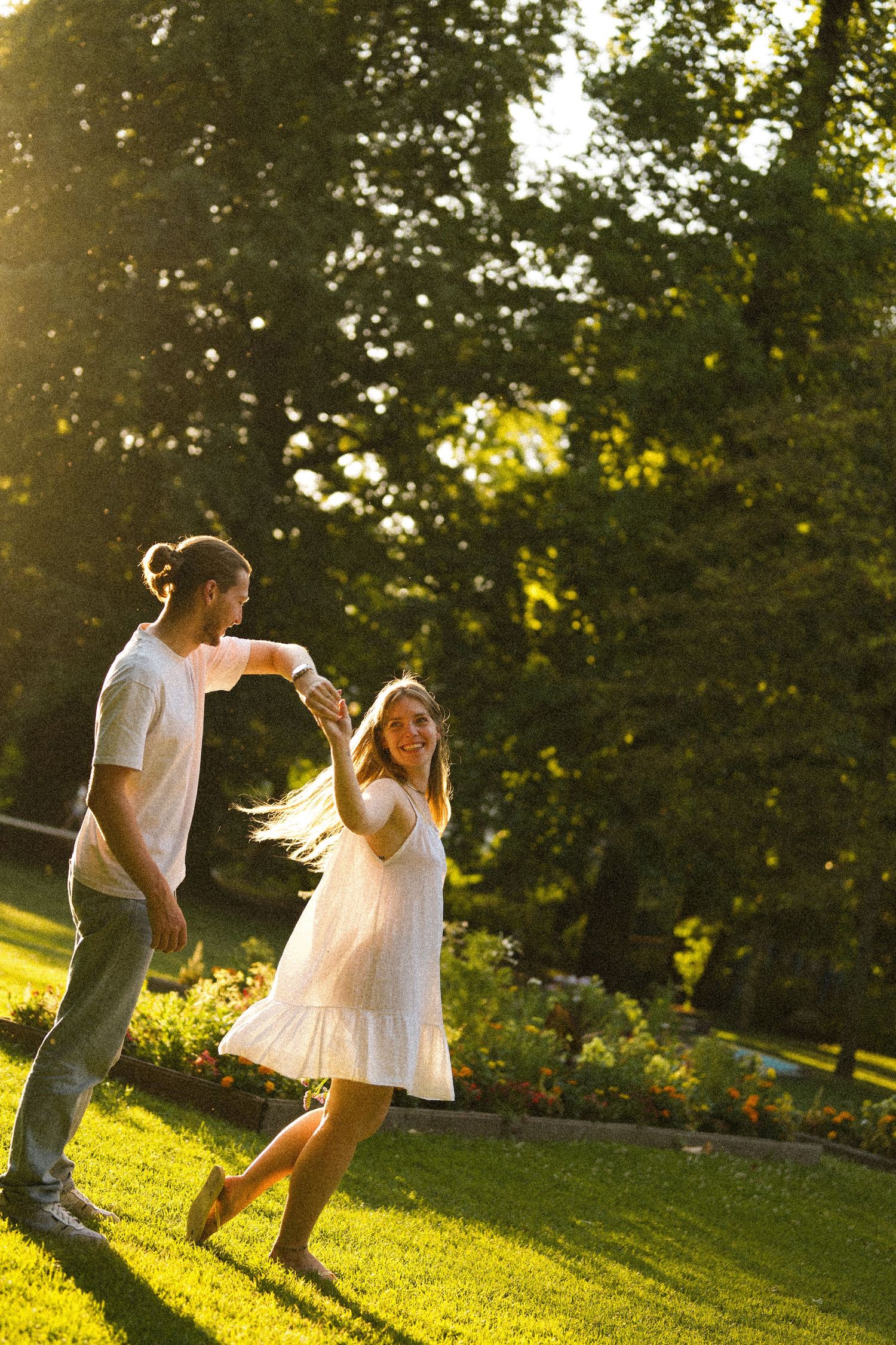 Joyful couple dancing and spinning on a green lawn in a park during a warm golden sunset.