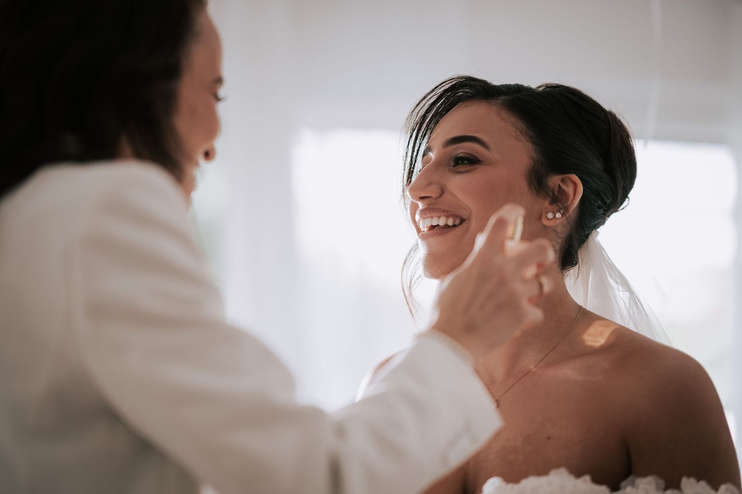 Smiling bride in white lace gown getting makeup touch-up before wedding ceremony.