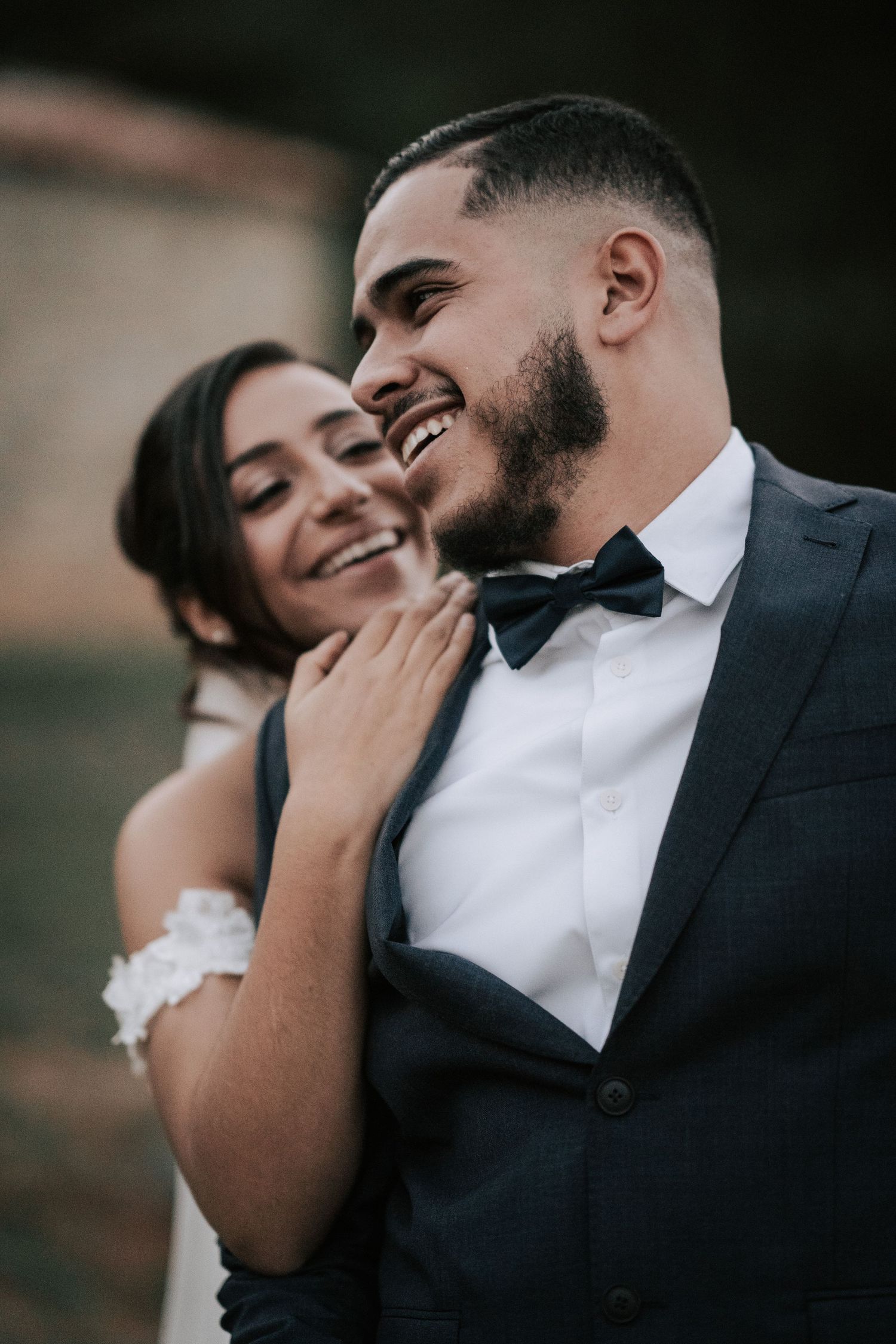 Smiling groom in a dark suit and bow tie embraced by his laughing bride on their wedding day.