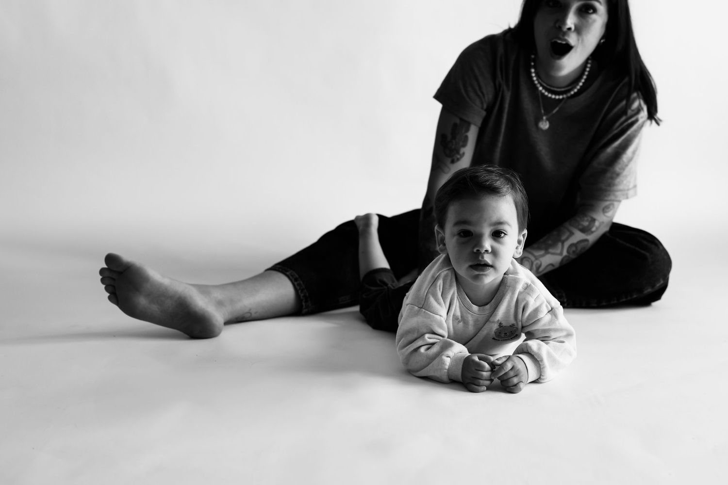 Black and white photo of baby lying in front of tattooed mother on white floor.