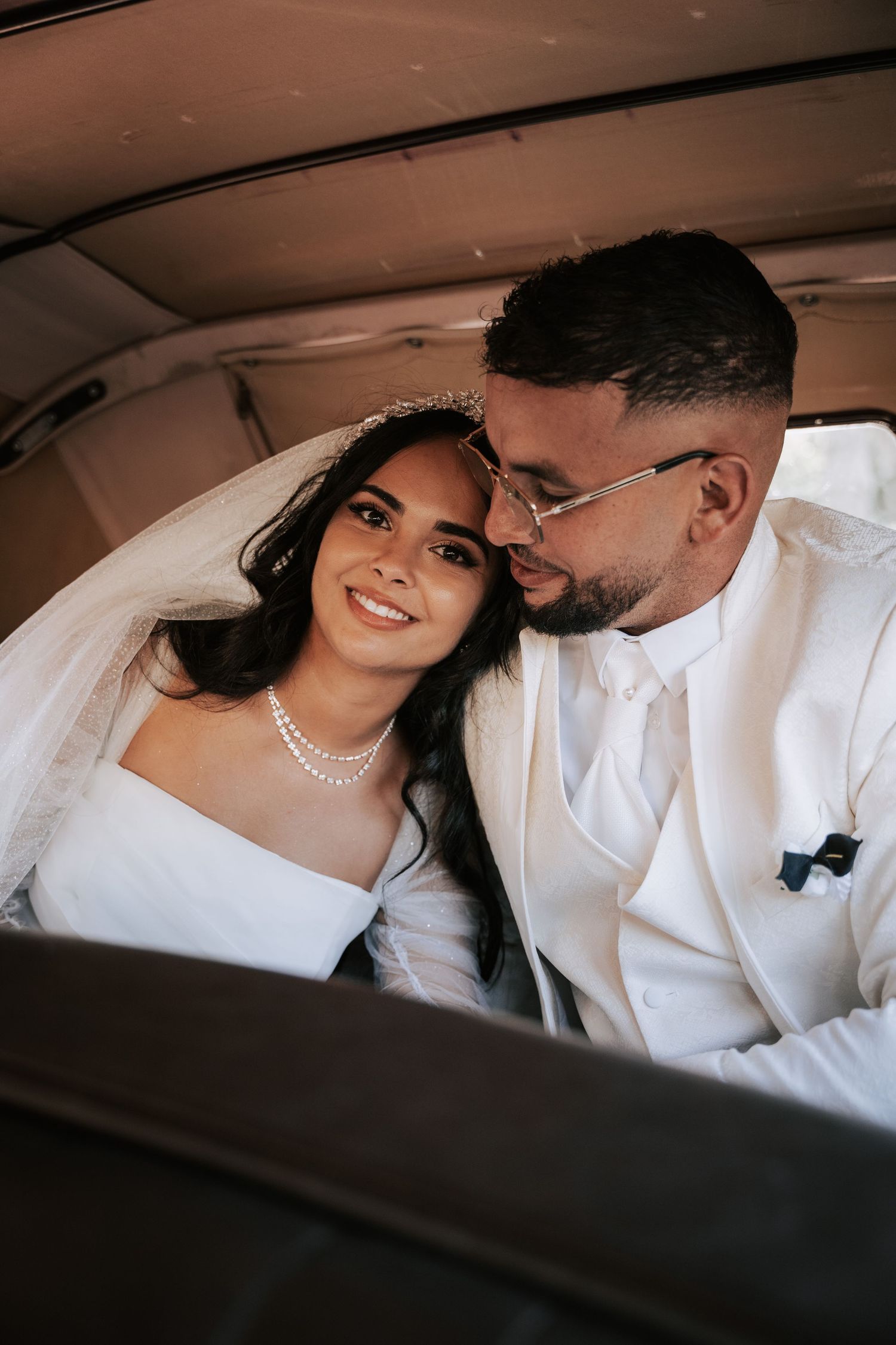 Bride and groom in white suit share tender moment together inside vintage car.