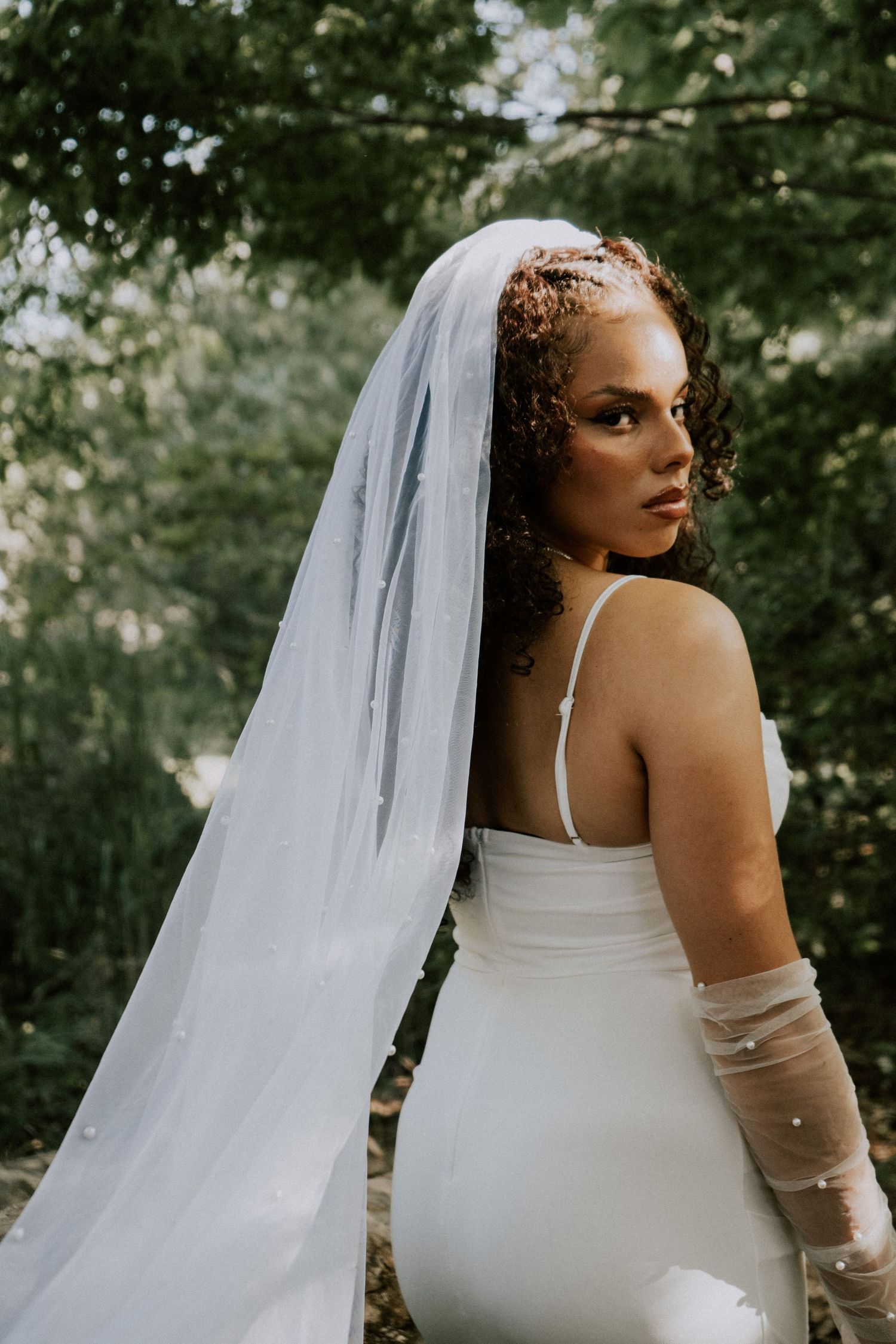 Bride in fitted white gown and long pearl veil looks over her shoulder in a lush green outdoor setting.