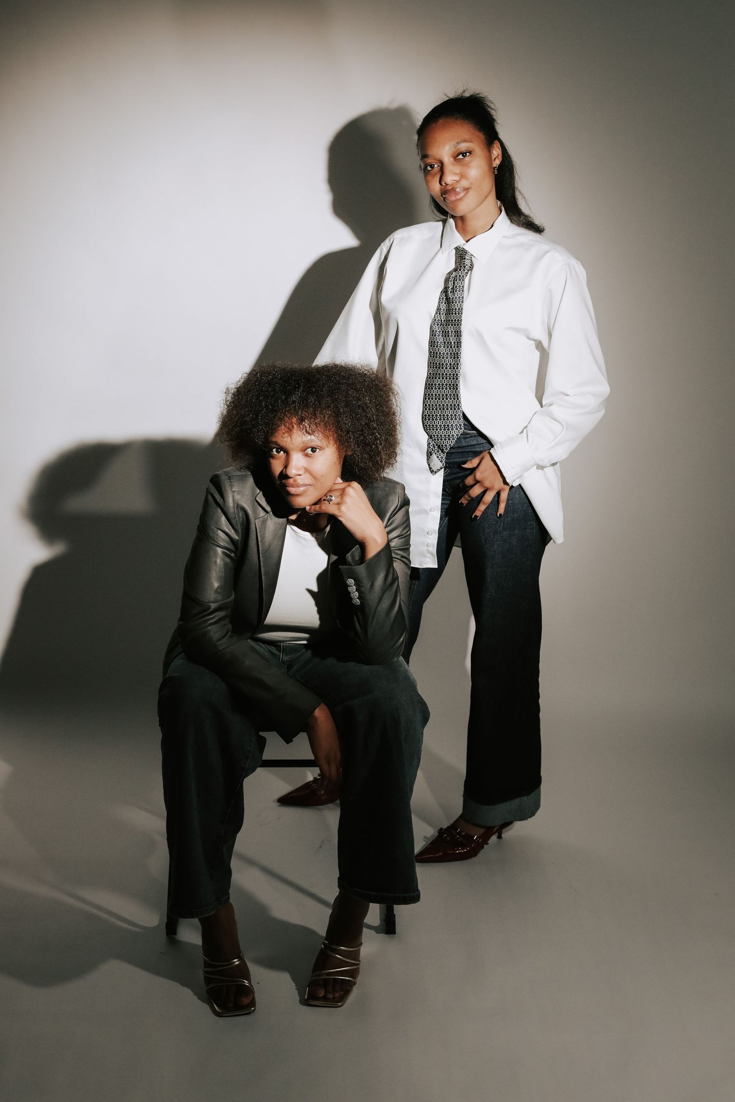 Two stylish women pose in a studio, one seated in leather jacket, one standing in white shirt and tie.