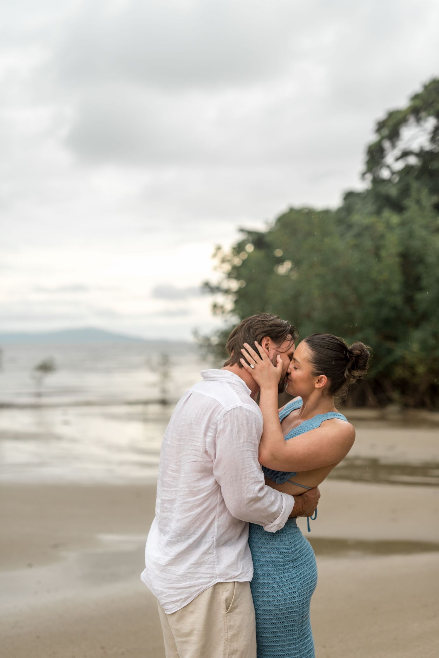 Romantic couple kissing on a beach with calm water and dense tropical trees in the background.