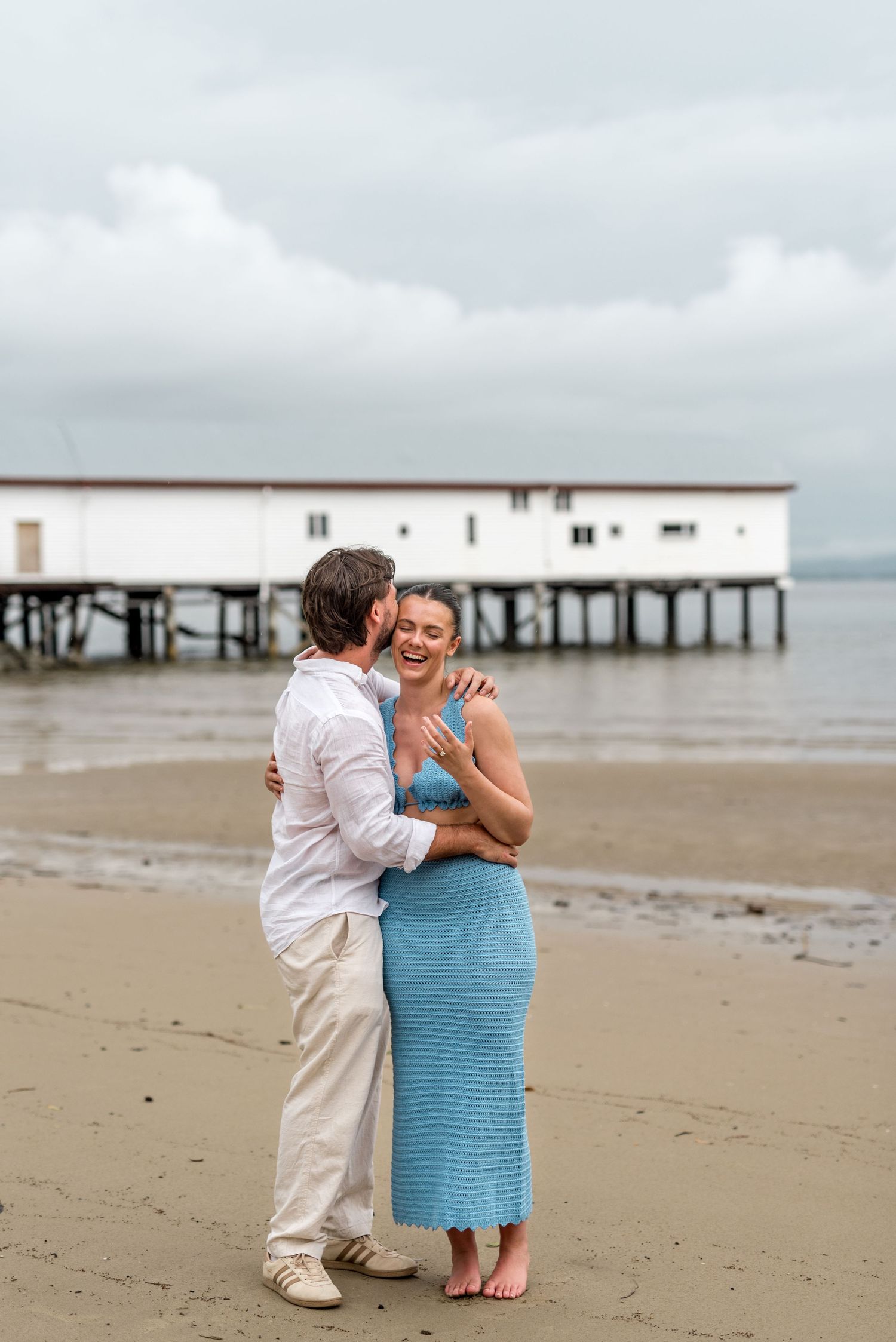 Happy couple laughing and embracing on a sandy beach with a white waterfront building on stilts in the background.