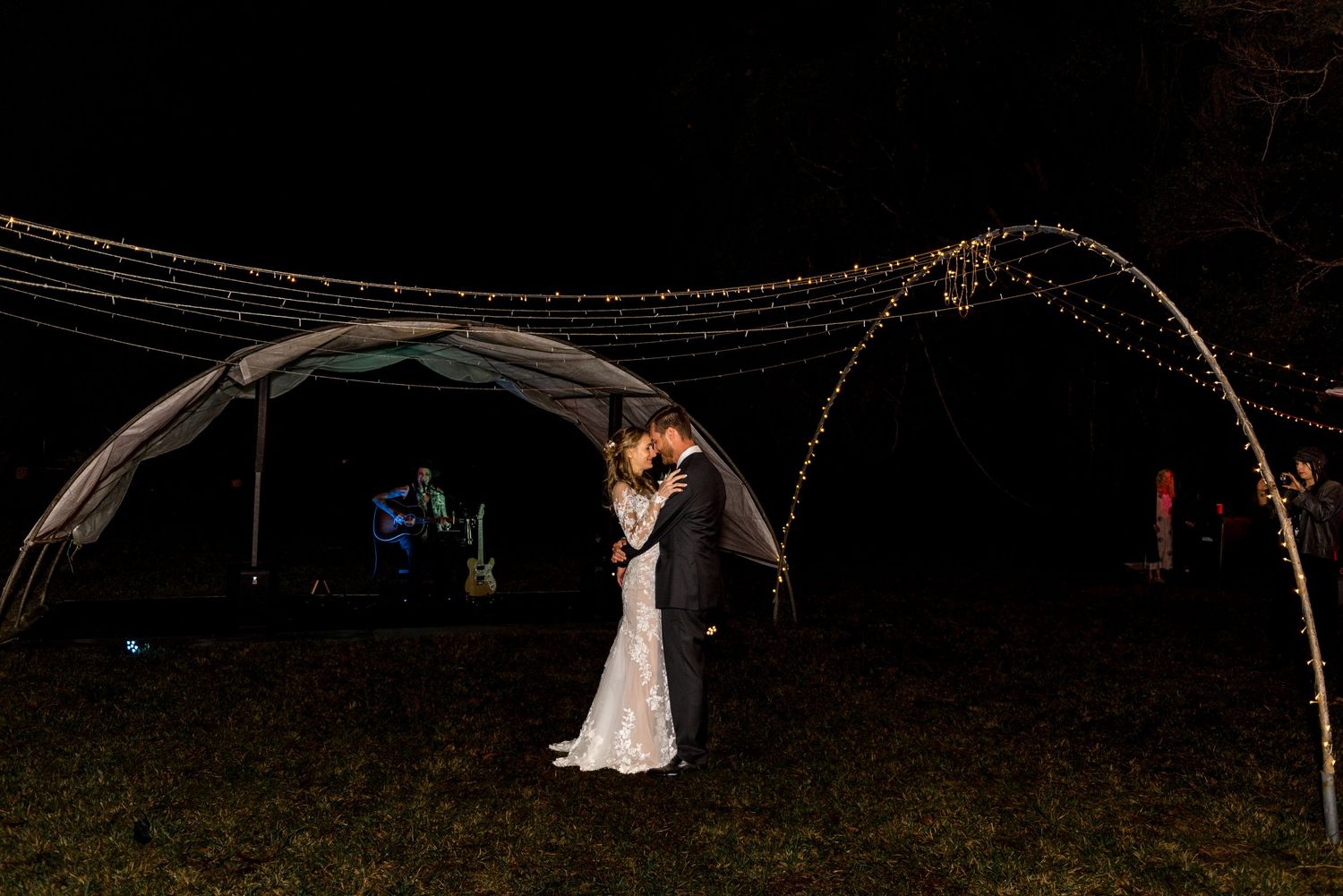 Bride and groom share a romantic kiss under an arch of twinkling fairy lights at their outdoor night wedding.