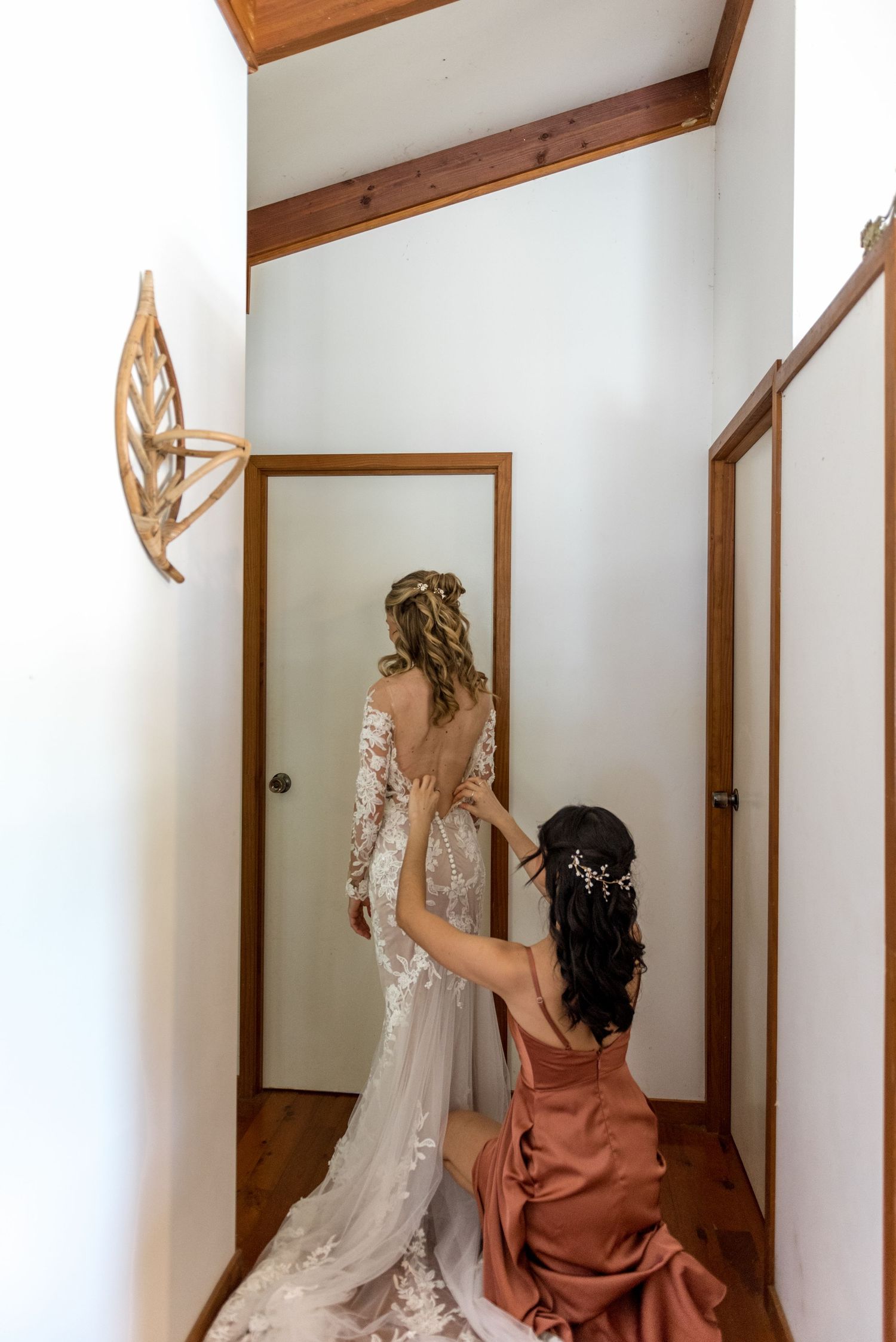 Bridesmaid in a rust dress helping the bride button her lace wedding gown in a hallway.
