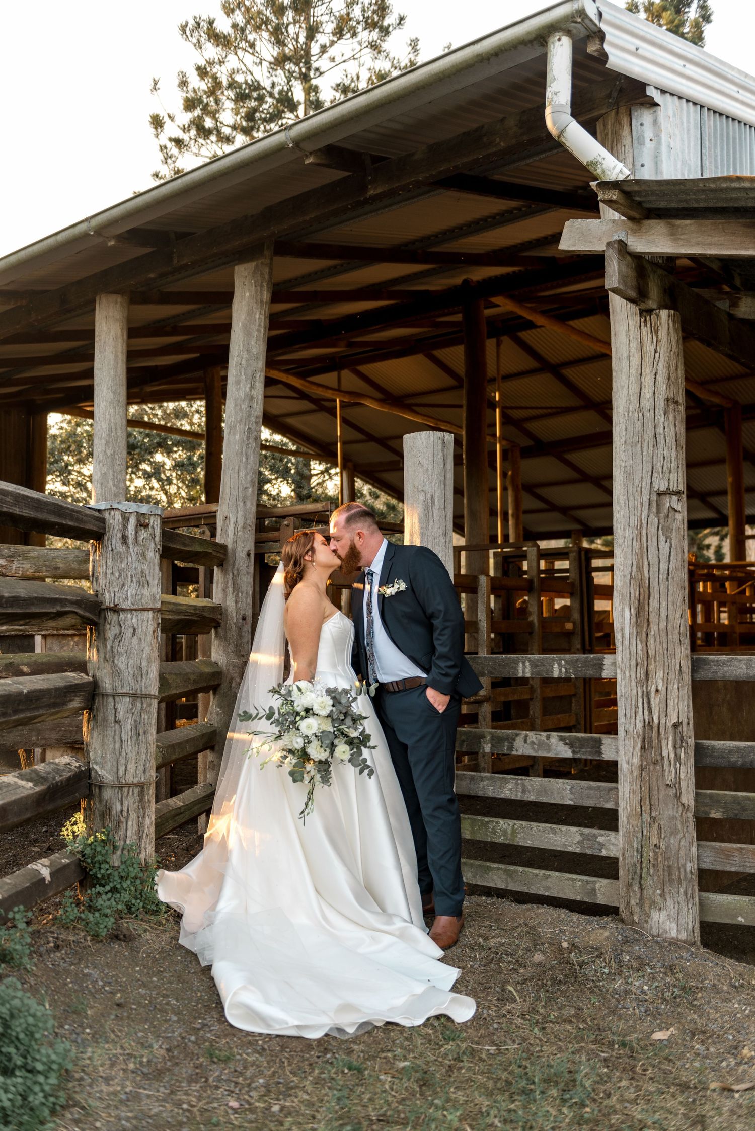 Bride and groom share a kiss under rustic wooden barn structure at golden hour.
