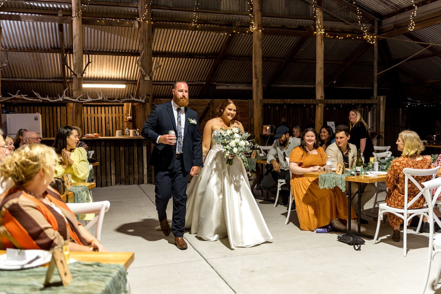 Bride and groom make grand entrance into rustic barn wedding reception with seated guests.