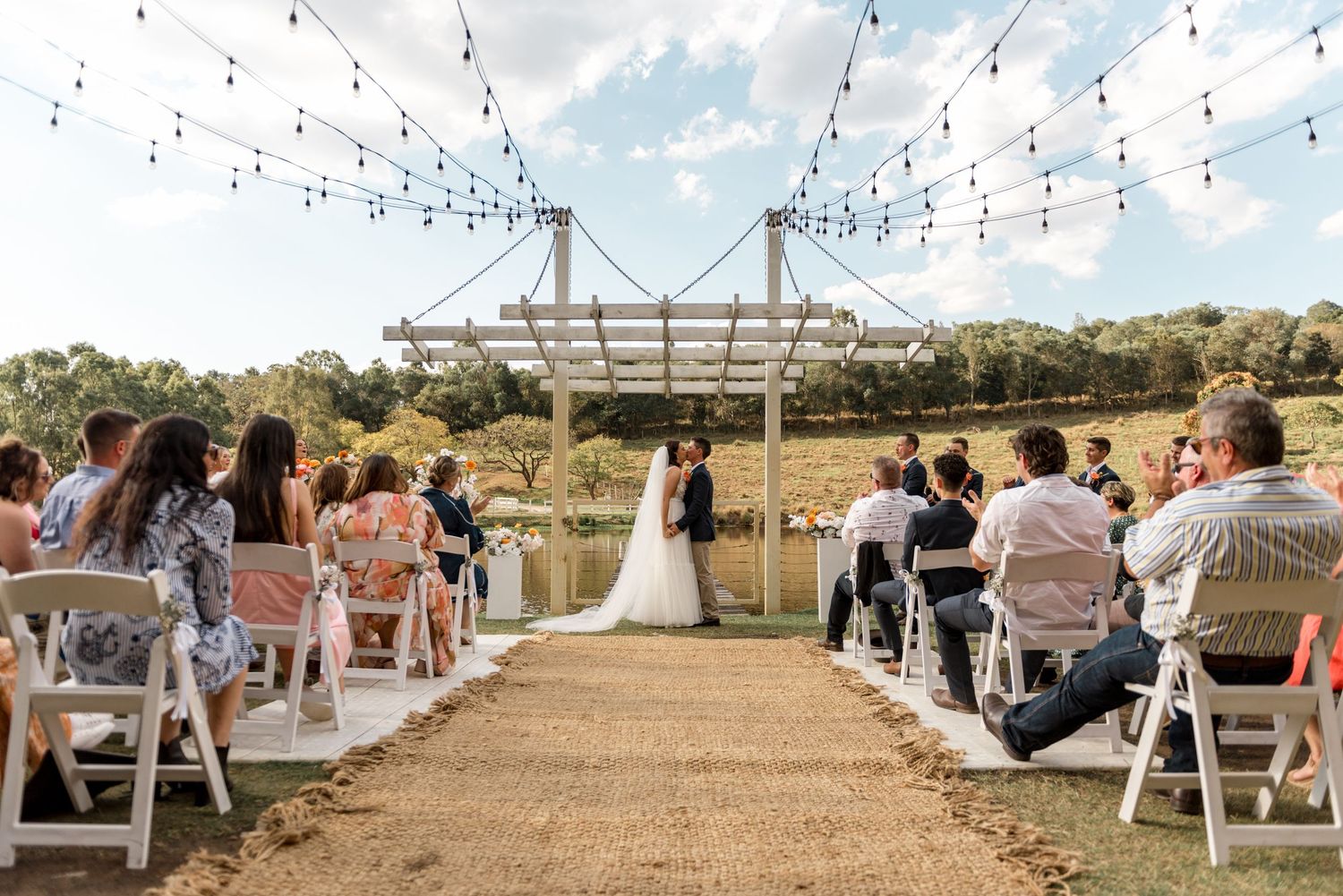 Bride and groom share a kiss at lakeside ceremony altar under string lights with seated wedding guests.