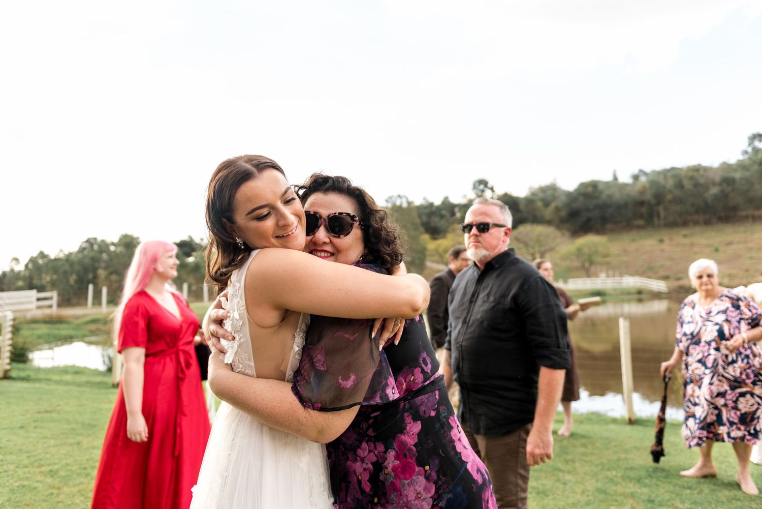 Bride in lace gown hugs a guest in floral dress at outdoor wedding reception near a scenic pond.