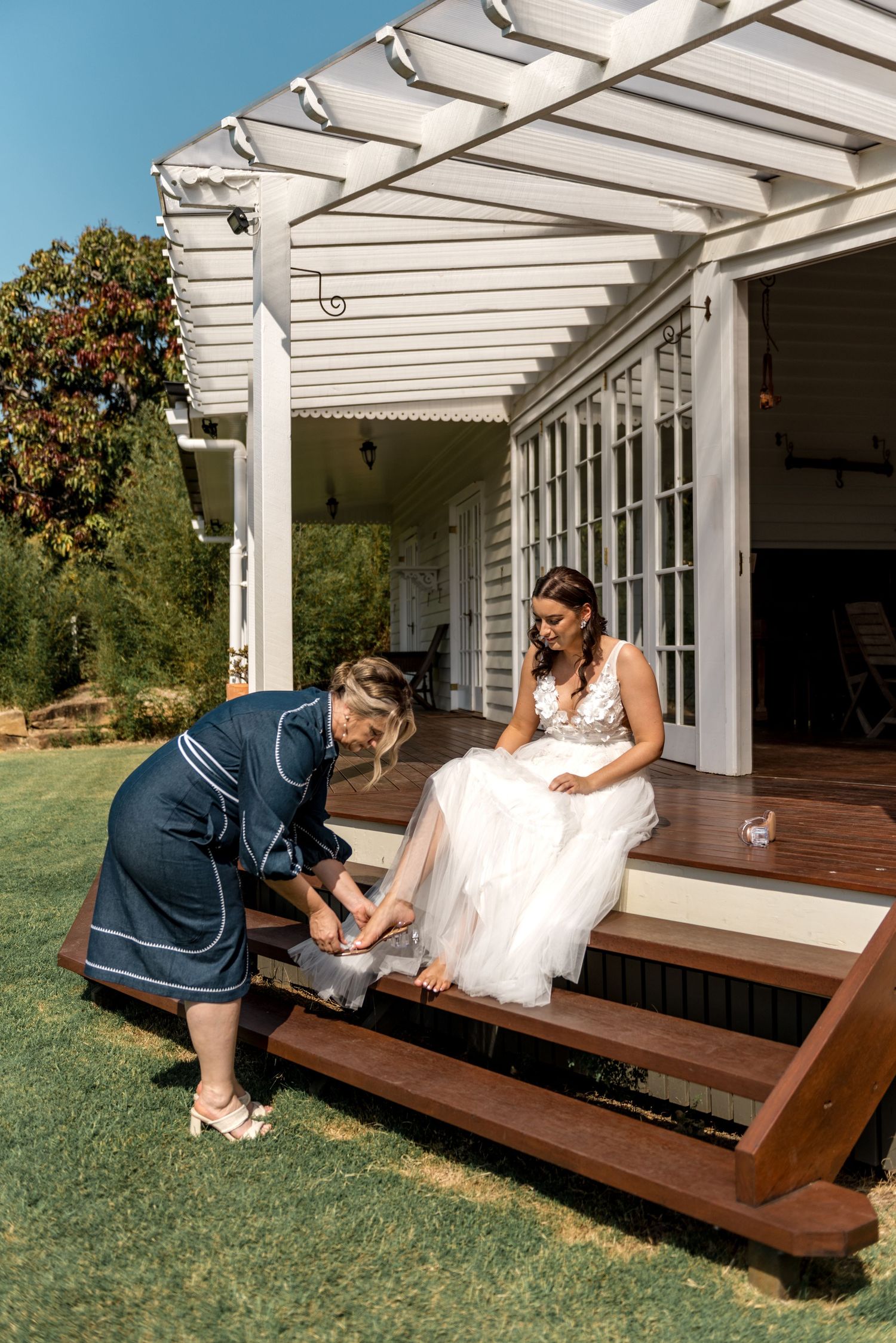 Woman in navy robe helping bride with shoes on timber steps of white weatherboard venue outdoors.