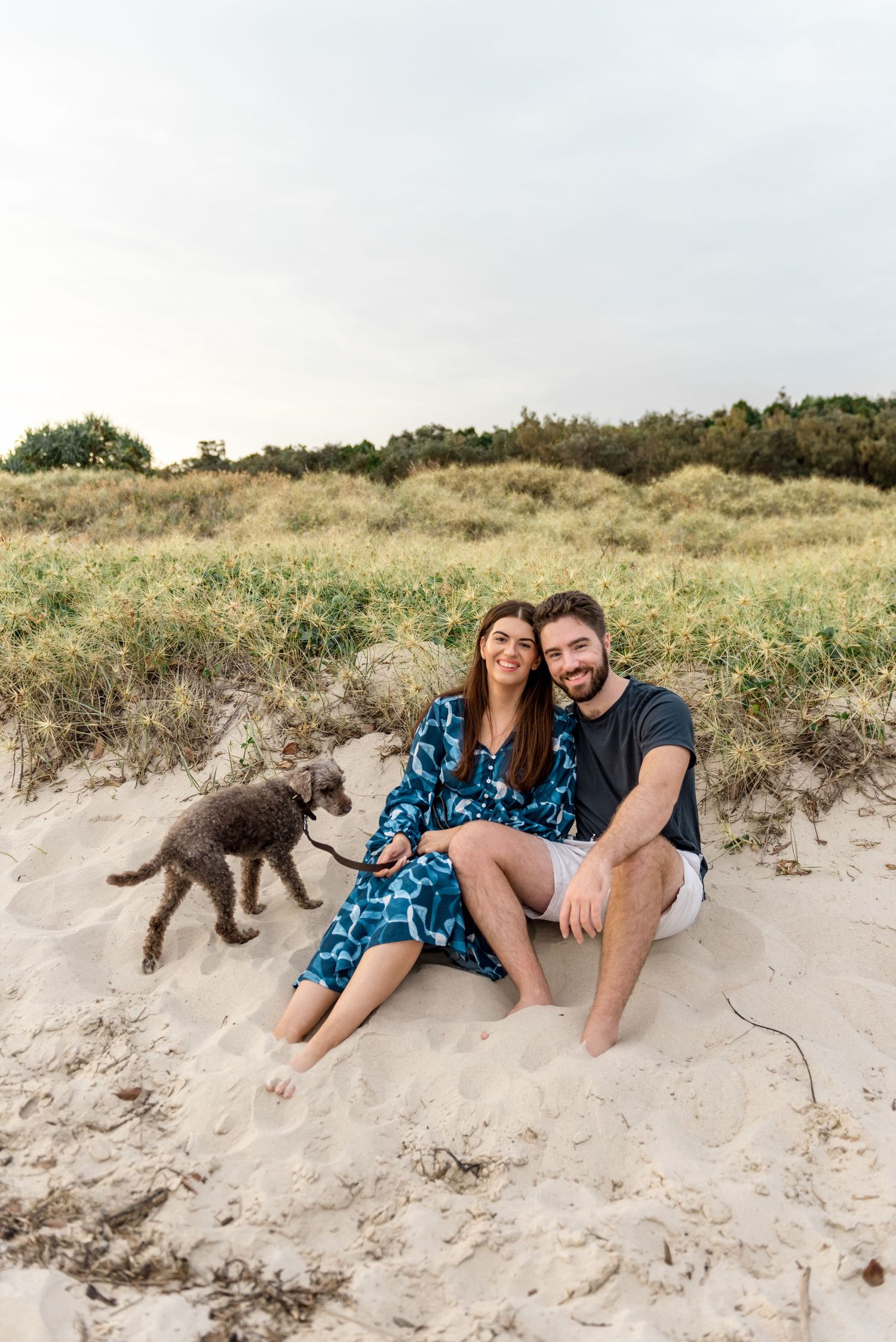 Smiling couple sitting on sandy beach dunes with gray dog on leash, beach grass behind them.