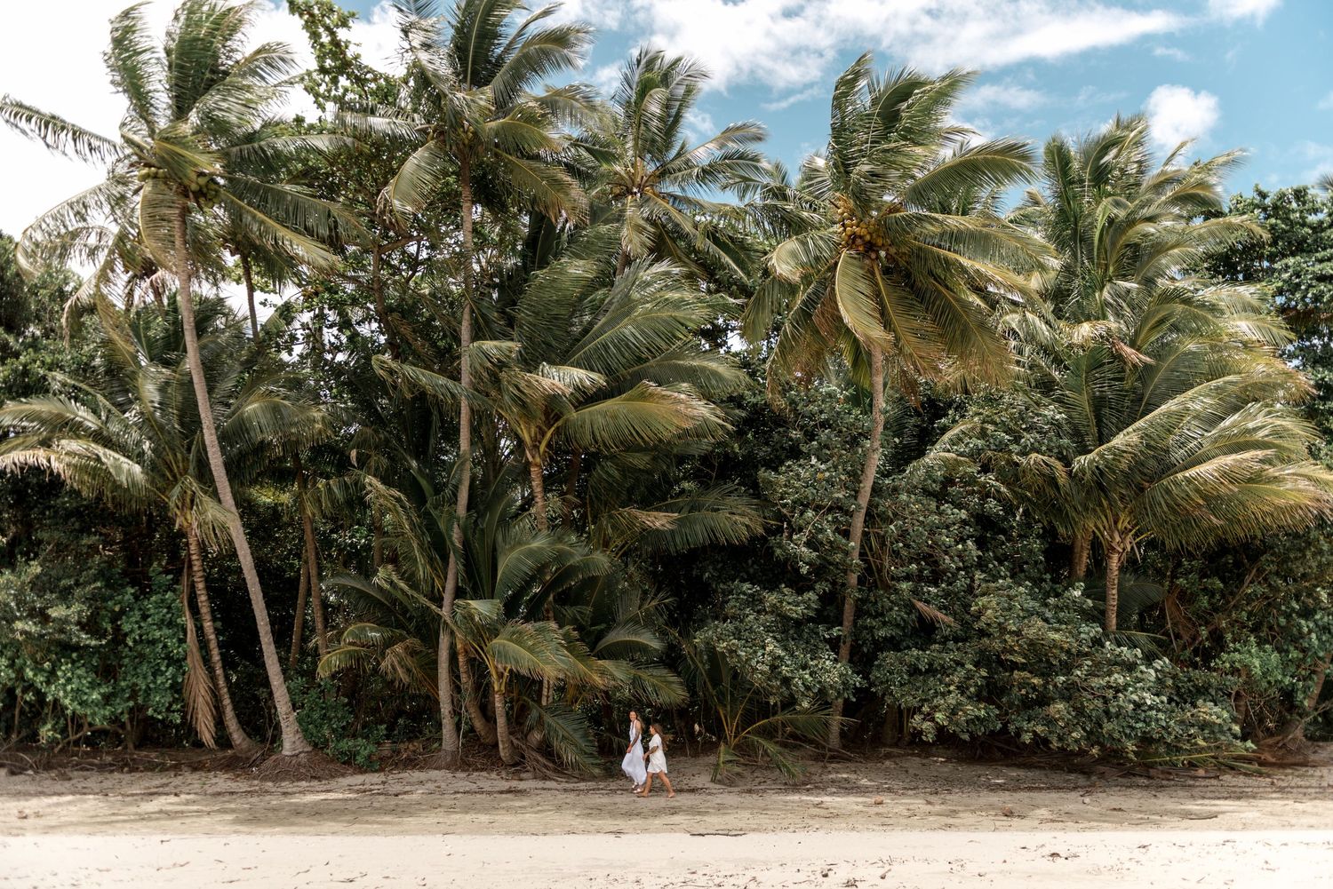 Two women in white dresses walking hand in hand on a sandy beach lined with tall tropical palm trees.