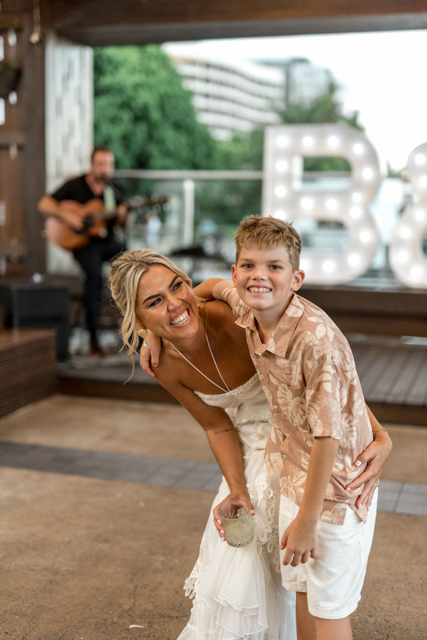 Smiling bride in white dress hugging young boy at outdoor wedding reception with guitarist performing in background.