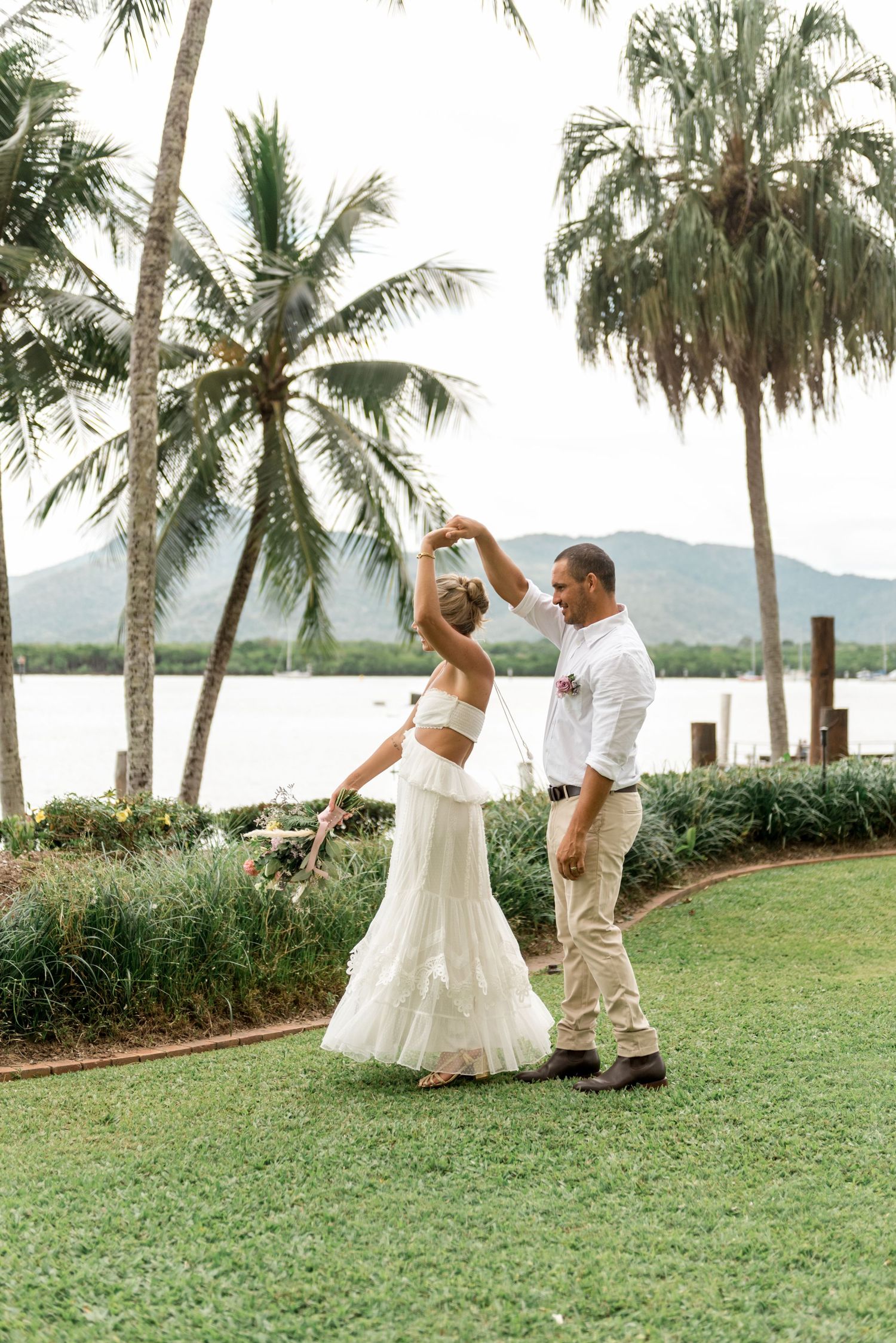Bride and groom dance joyfully on green lawn with tropical palm trees and mountain lake backdrop.