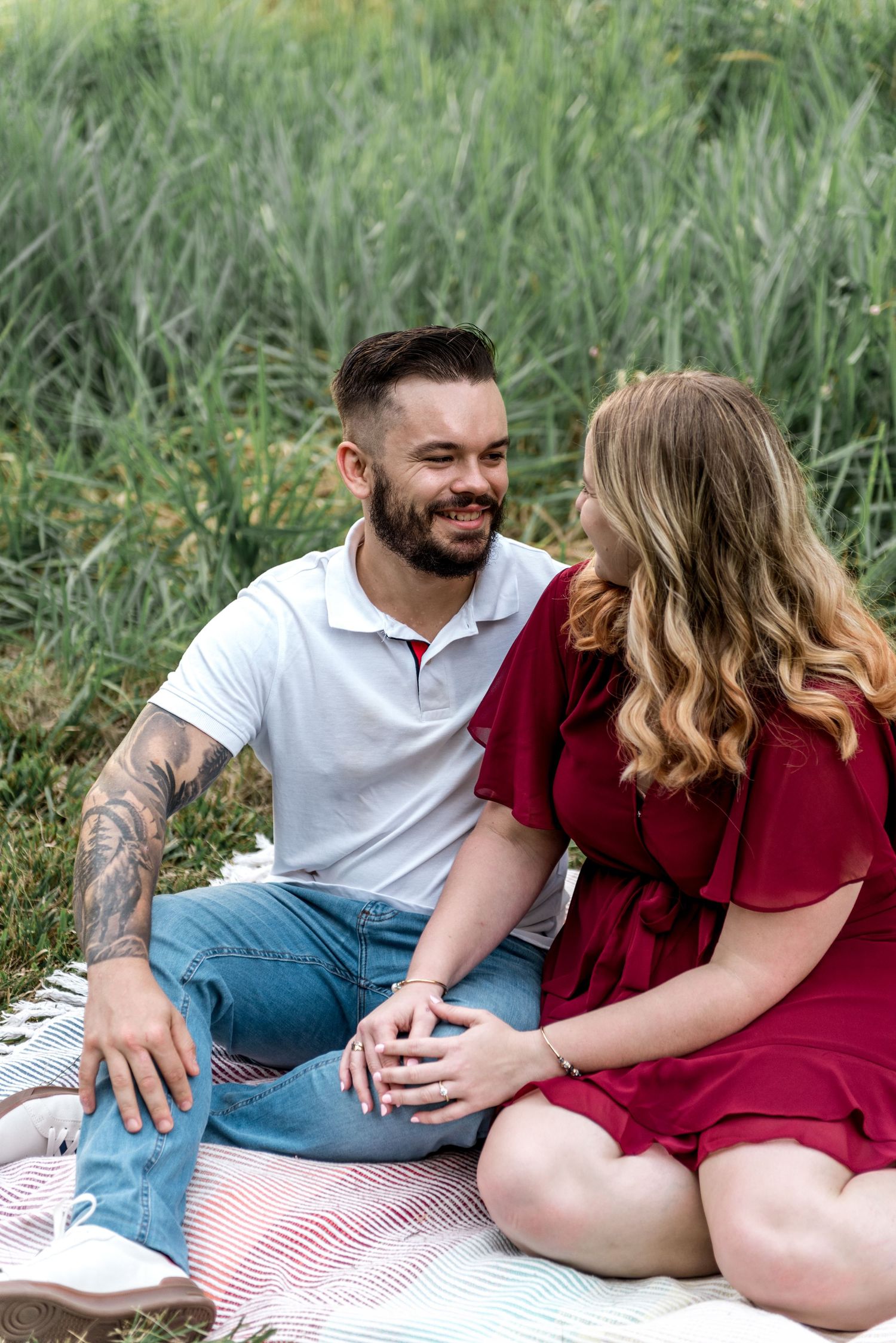 Tattooed man in white polo smiling at blonde woman in red dress while sitting on picnic blanket outdoors.