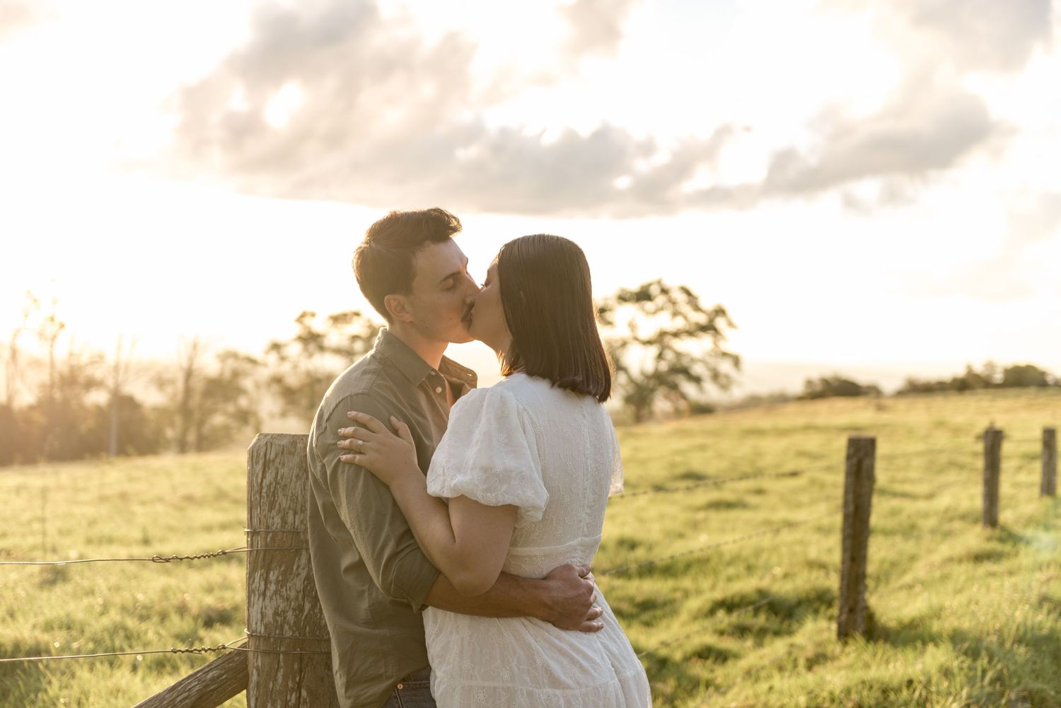 Couple sharing a kiss at sunset beside a wooden fence in a scenic green pasture.