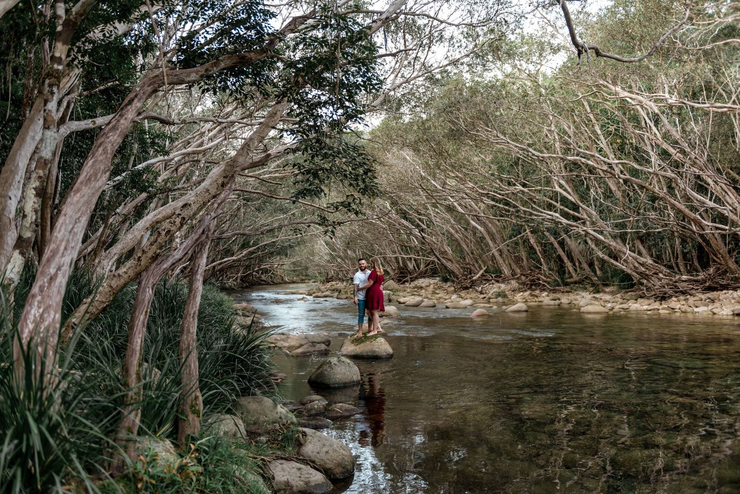 Couple standing on rocks in a peaceful forest river surrounded by twisted trees and lush greenery.