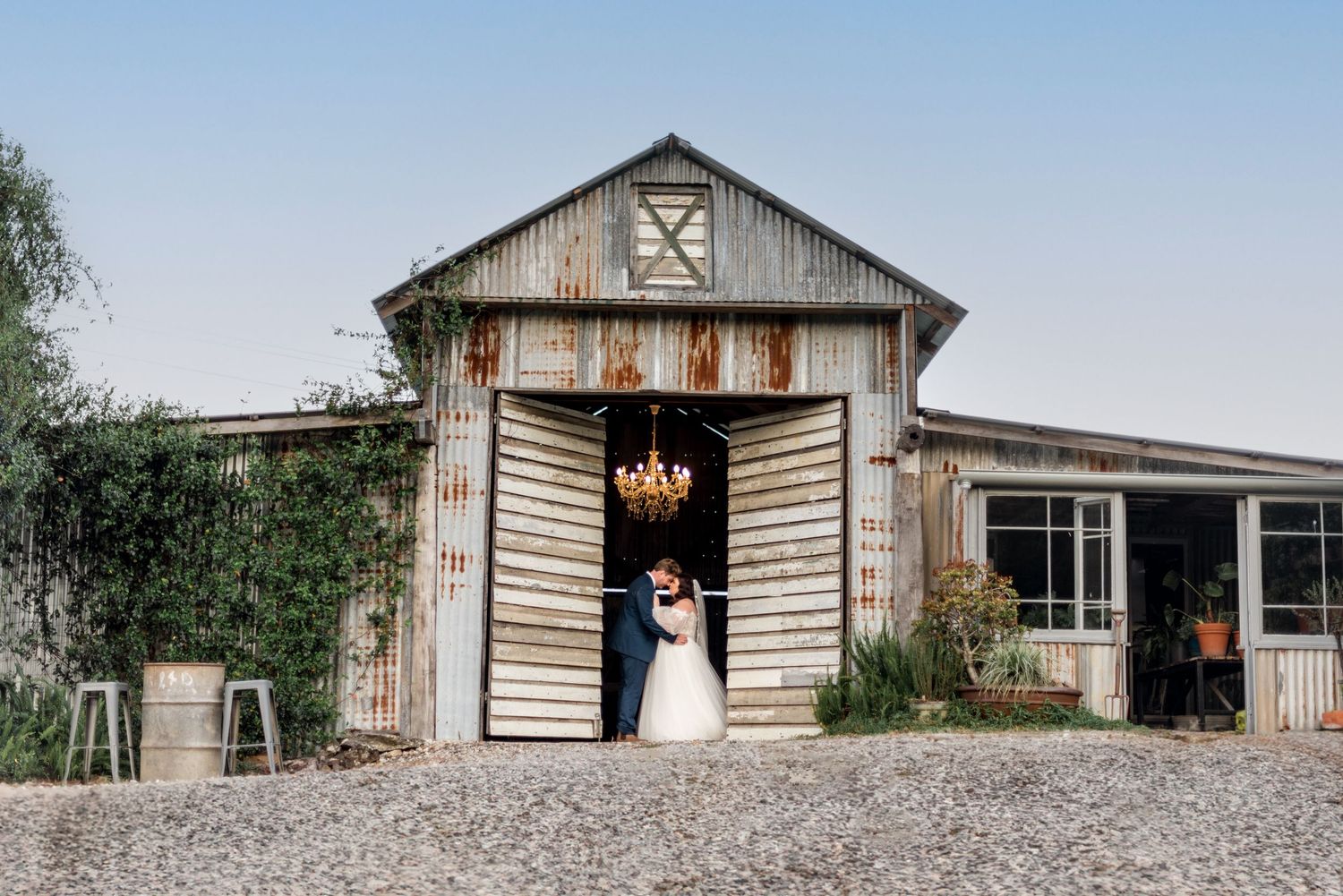 Bride and groom share a kiss in the doorway of a rustic barn venue with a chandelier overhead.