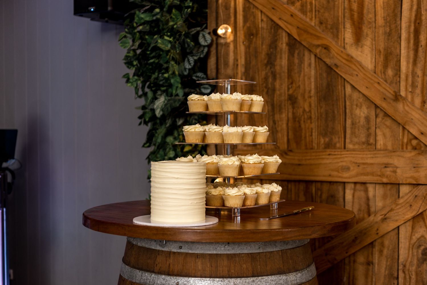 Elegant wedding cake and cupcake tower displayed on a wooden barrel in front of rustic barn doors.