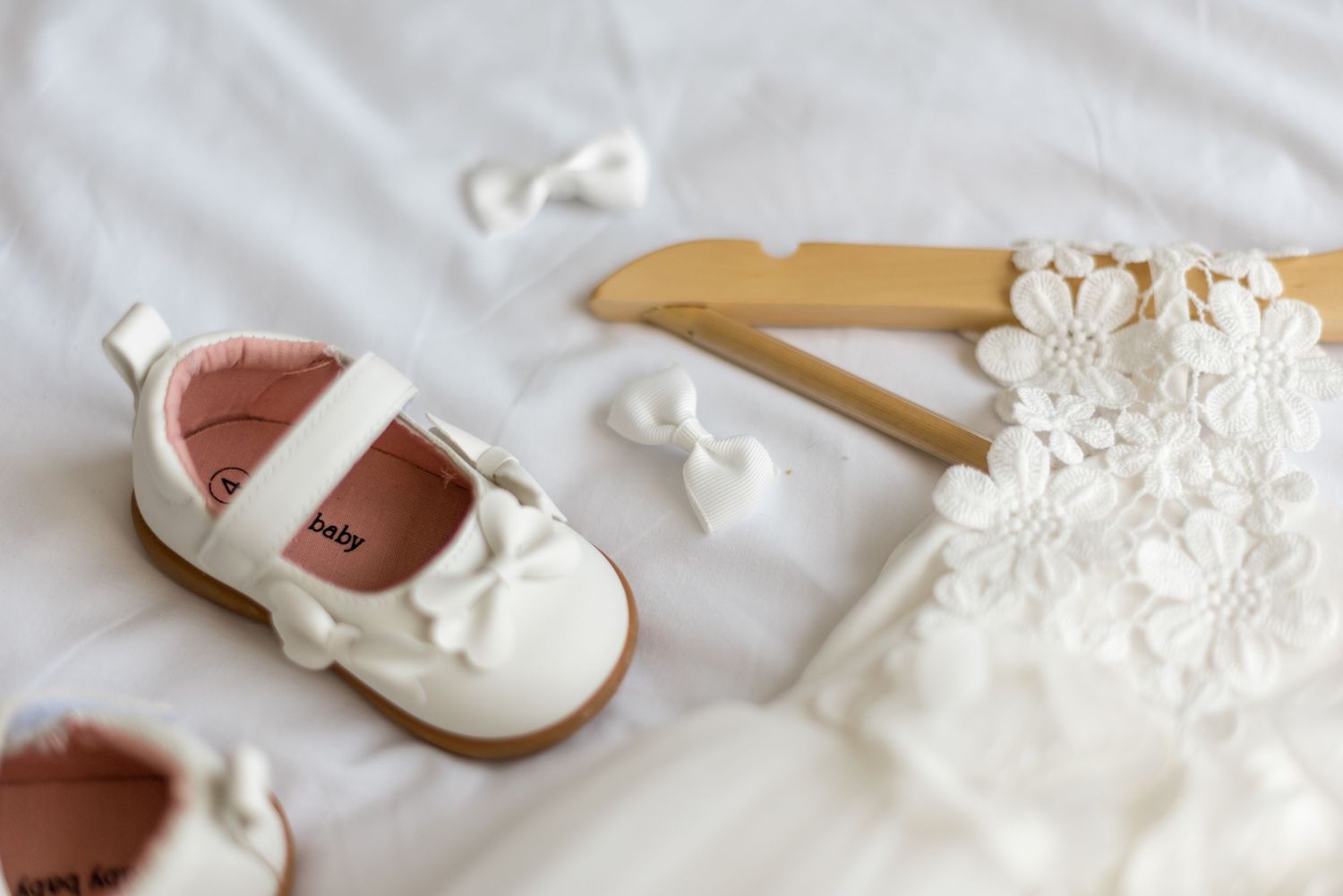 White baby Mary Jane shoes with bow next to lace flower girl dress on wooden hanger.