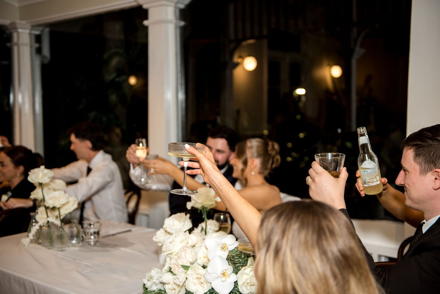 Wedding guests raising glasses in a toast at a romantic evening reception with white roses.