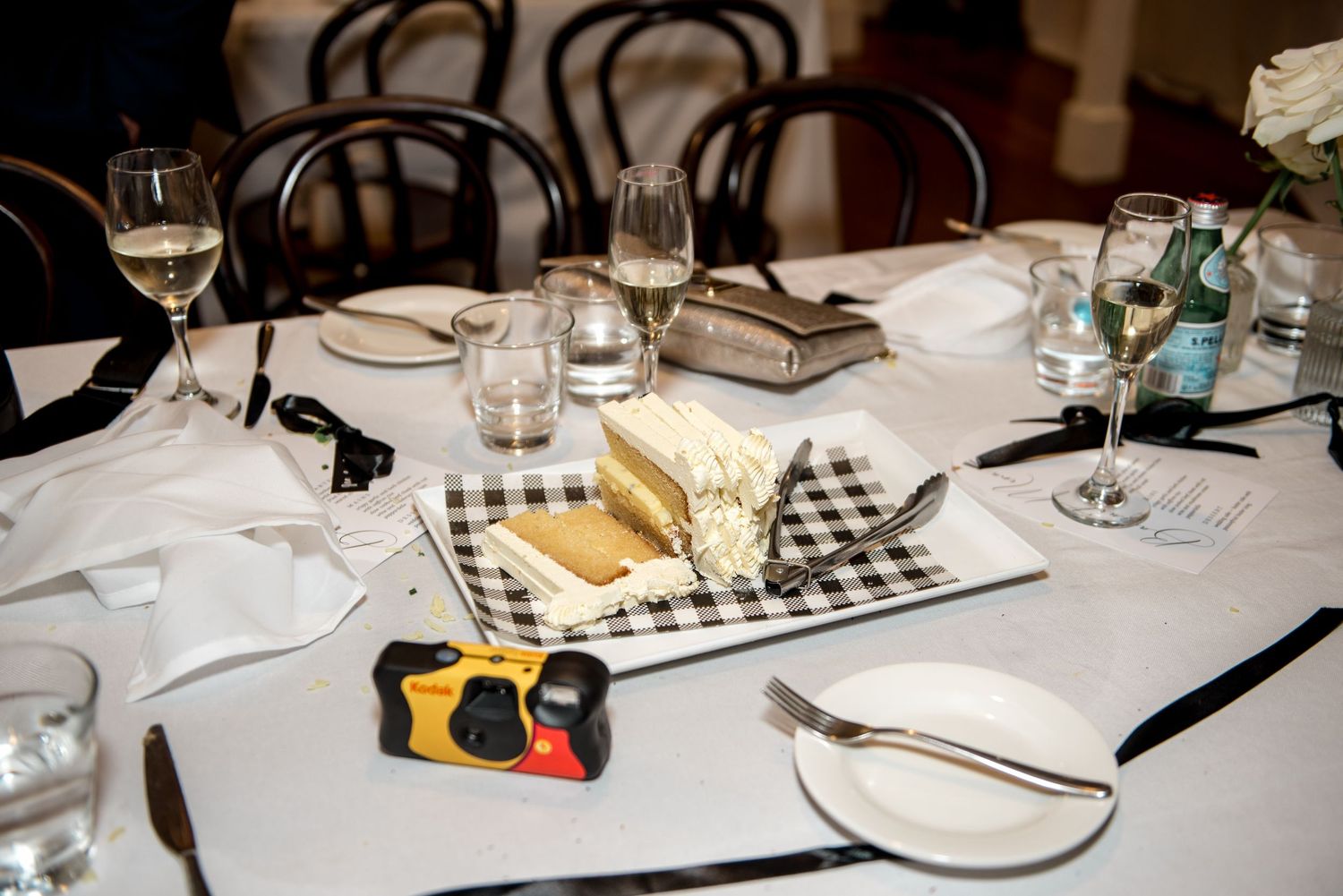 Elegant wedding reception table set with white linen, glassware, disposable camera, and gingham-wrapped favors.