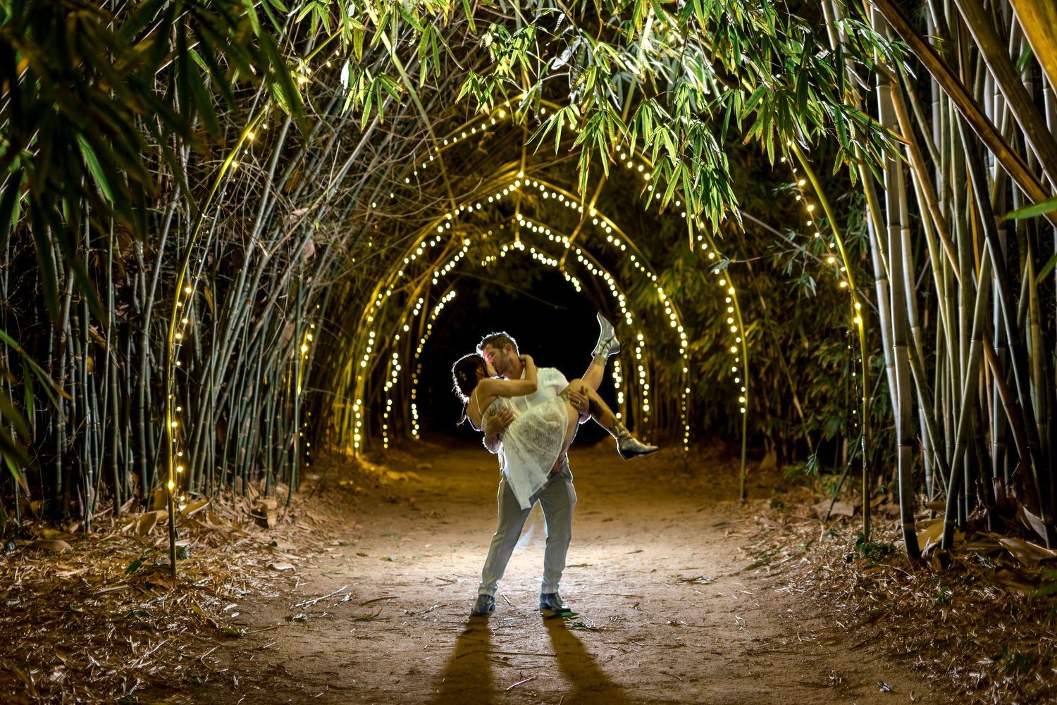 Groom lifting bride in a stunning bamboo tunnel decorated with fairy lights at night.
