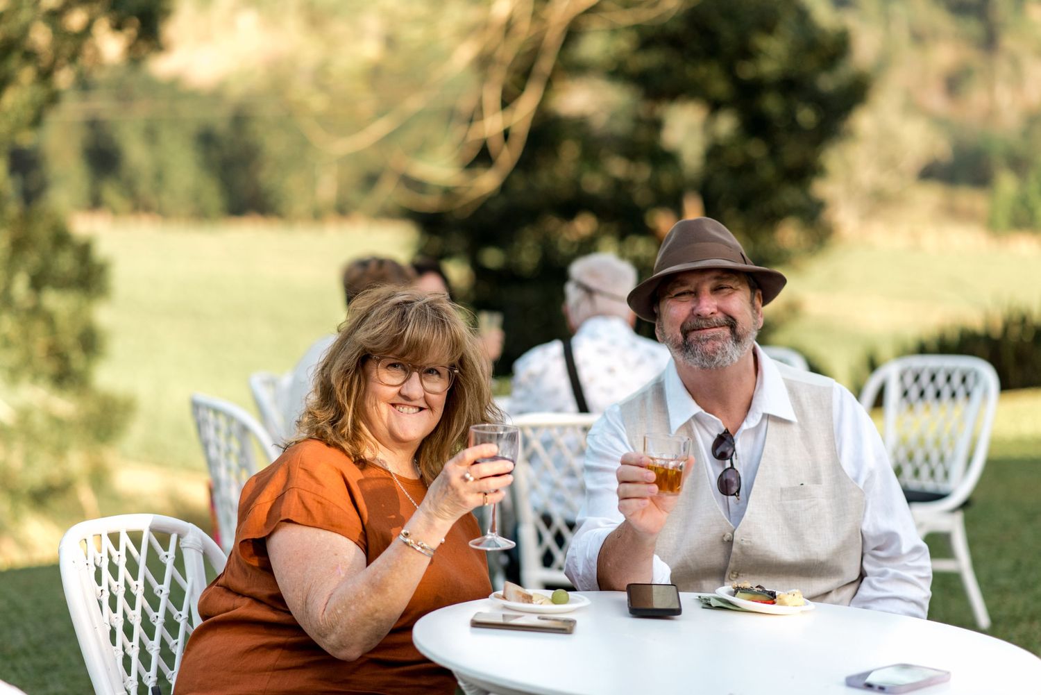 Couple smiling and raising glasses at outdoor winery table surrounded by lush green gardens.