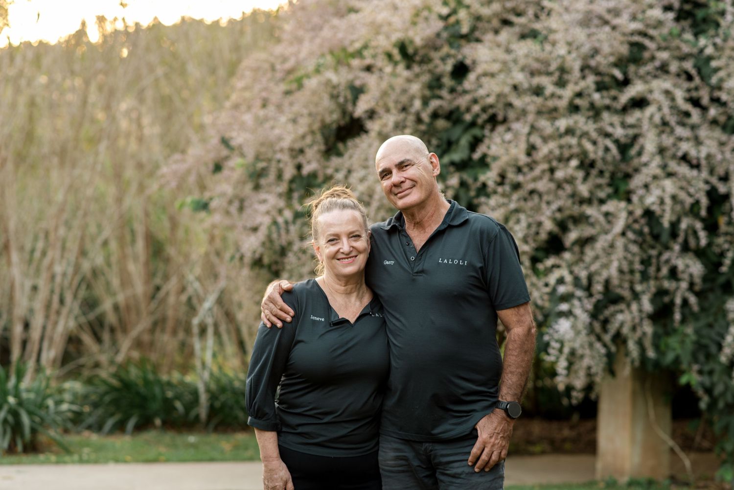 Smiling older couple embracing outdoors in front of flowering pink trees at dusk.