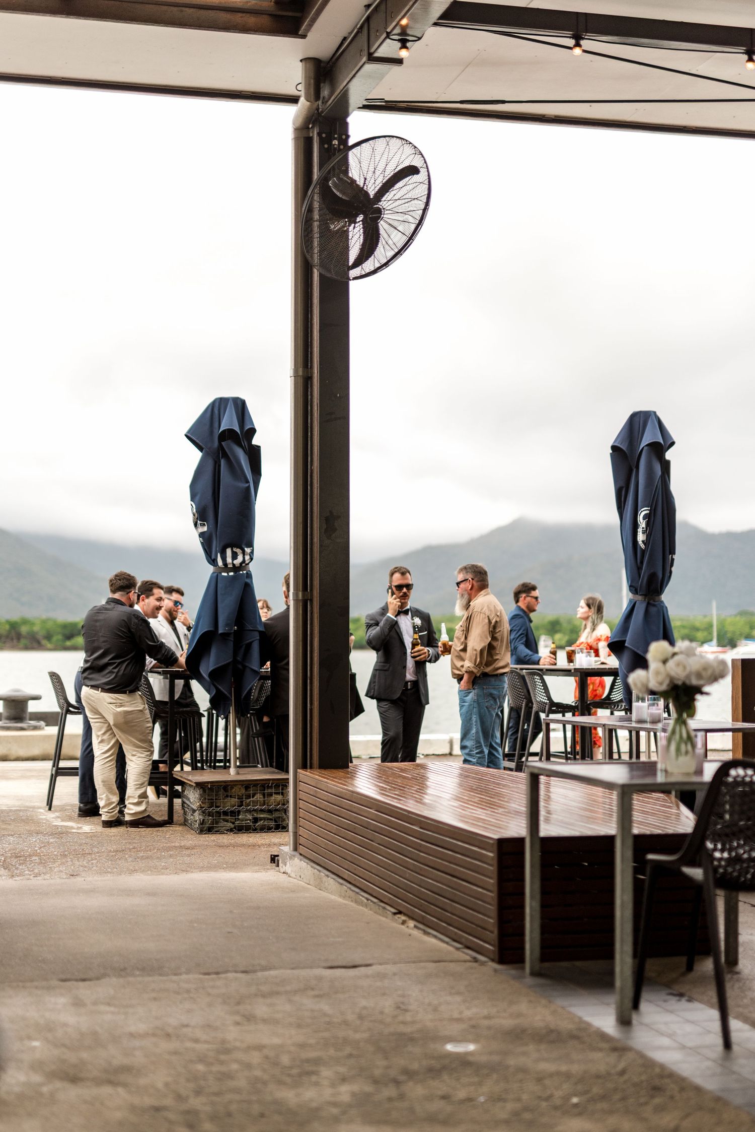 Wedding guests socializing on a waterfront venue terrace with navy umbrellas and misty mountain views.