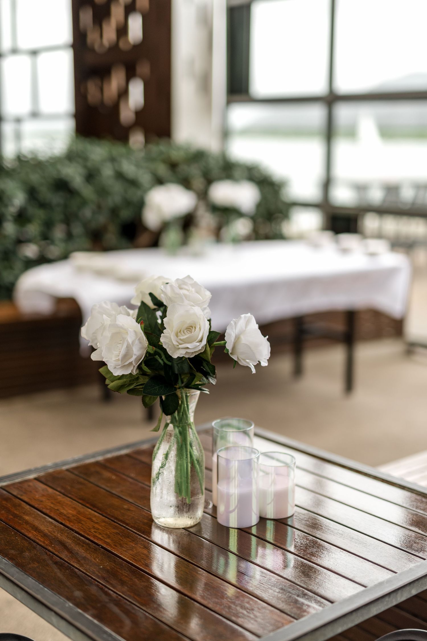 White roses in glass vase with candle on wooden table in elegant wedding venue.