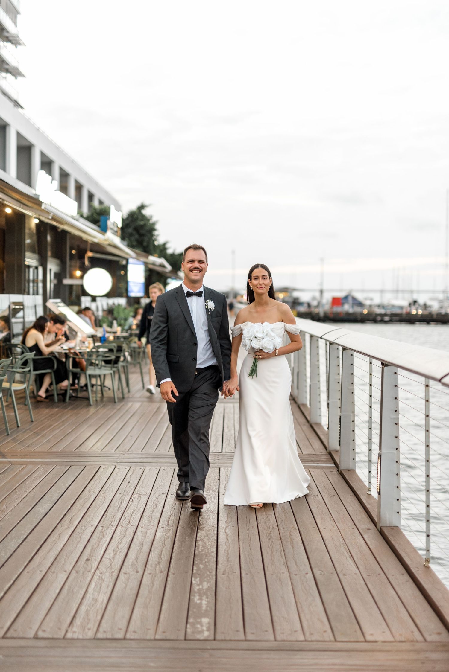 Bride and groom walking hand-in-hand along a waterfront wooden boardwalk, bride in off-shoulder gown holding white bouquet.