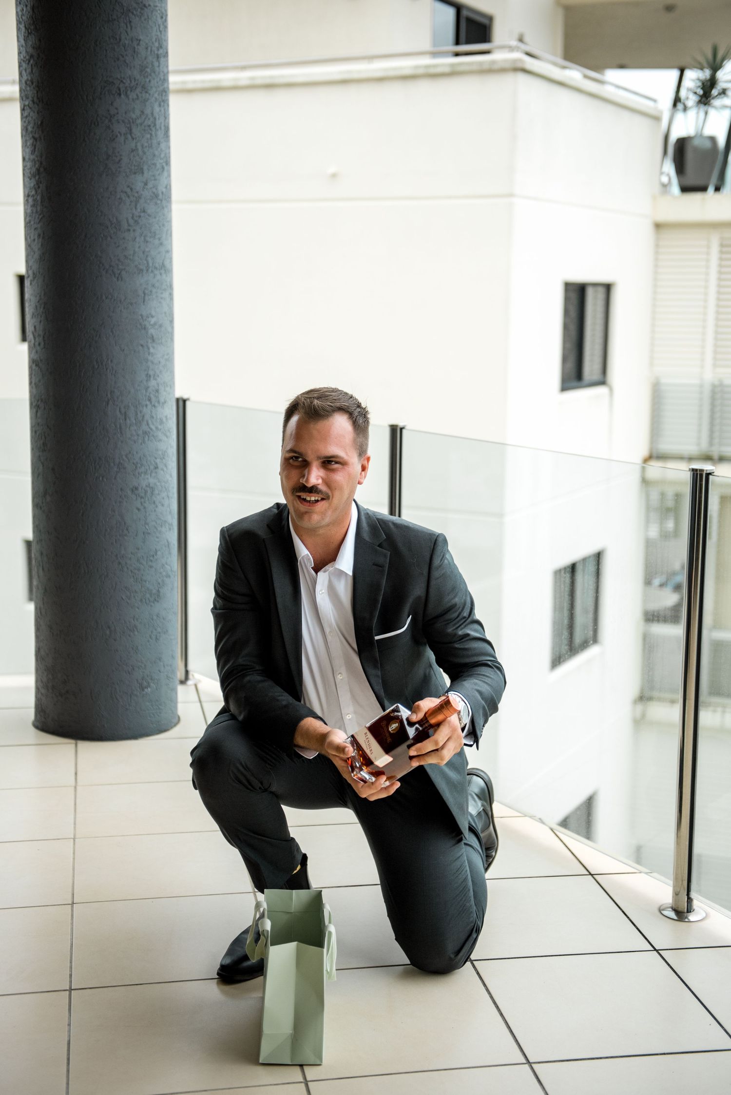 Groom in dark suit kneels on balcony holding phone and gift box, smiling on wedding morning.