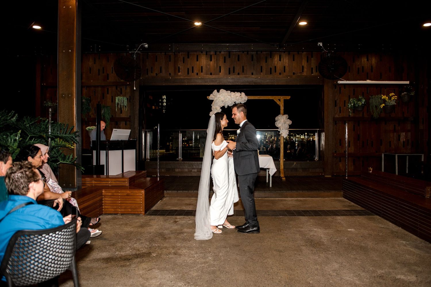 Bride and groom share their first dance indoors beneath a white floral balloon arch at their evening wedding reception.