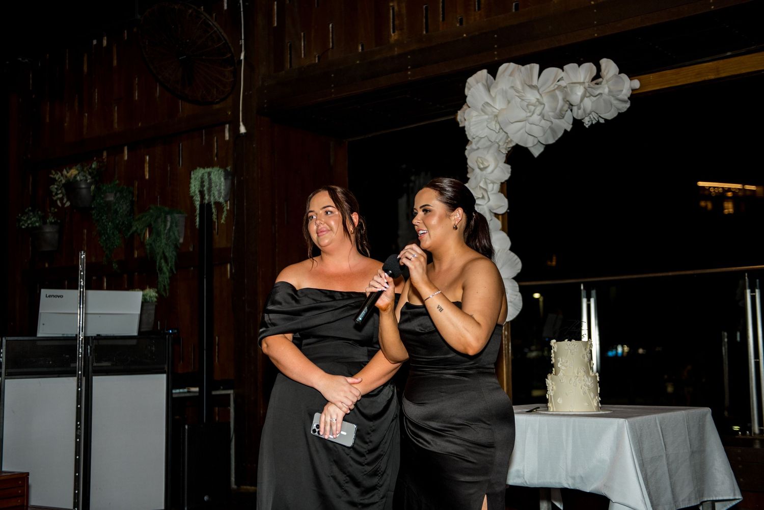Two women in black dresses give a wedding speech together, standing near a white cake with balloon arch decoration.