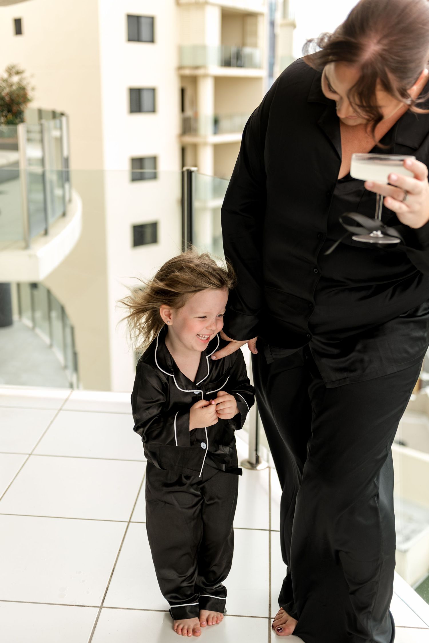 A woman in black satin pajamas holds a champagne glass while a young girl in matching black pajamas laughs on a balcony.