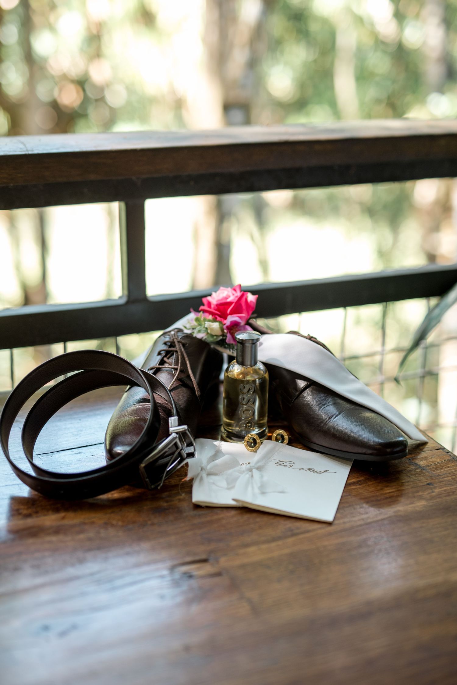 Wedding flat lay featuring brown leather shoes, belt, cologne, boutonniere, and vow cards on wood.