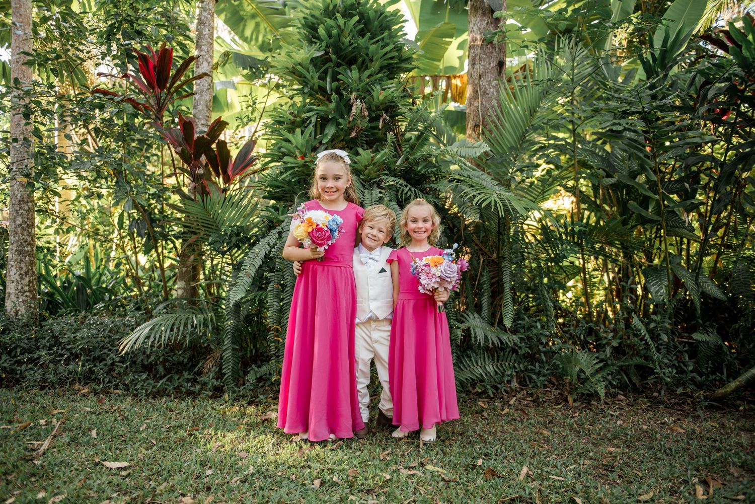 Two flower girls in hot pink dresses and a ring bearer in cream suit holding colorful bouquets in a lush tropical garden.