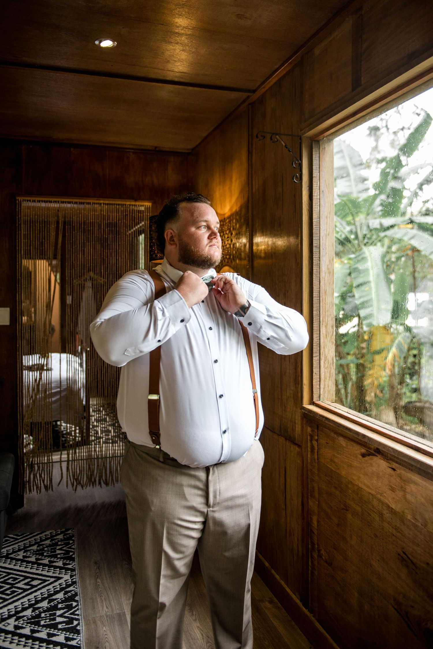 Groom adjusting his bow tie and suspenders while getting ready beside a window with tropical views.