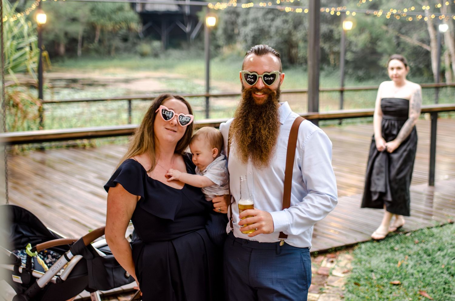 Couple with baby poses smiling at outdoor wedding reception with string lights and guests in background.