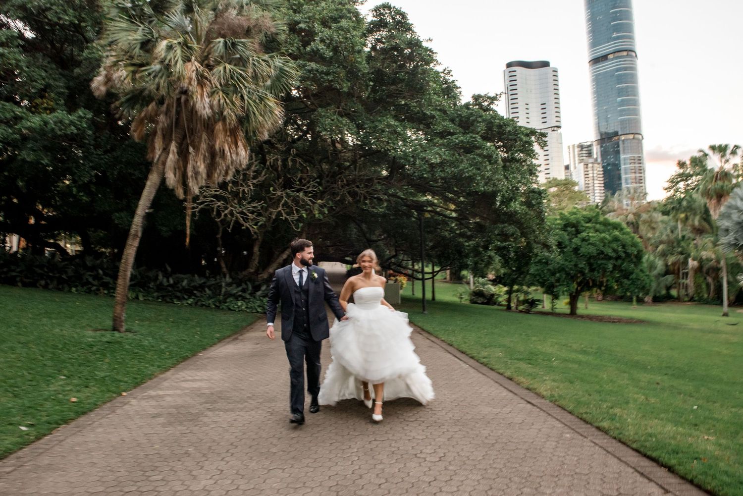 Bride in tiered tulle gown and groom walk hand-in-hand through a lush park with city skyline.